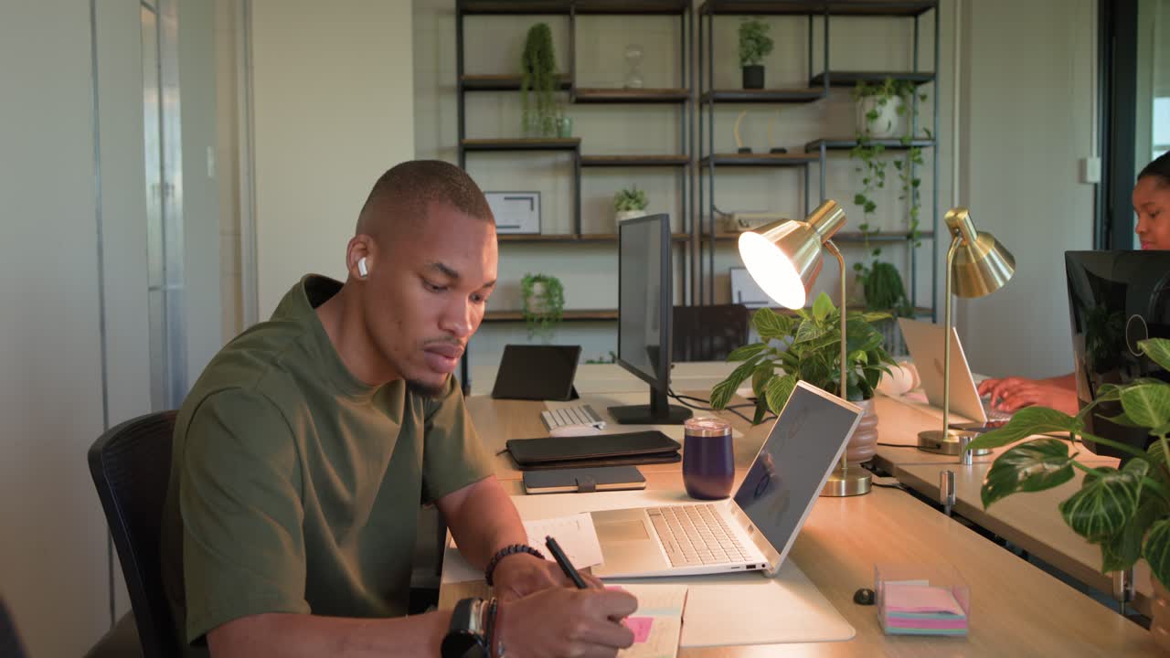 African man sorting notes, placing pink sticky notes, typing on laptop at desk after woman entering