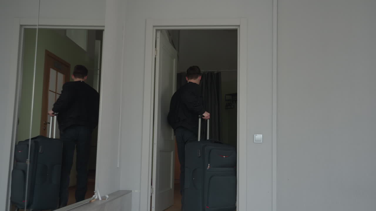 Sport man seen from behind enters apartment wearing black jacket after returning from sport trip, facing closed white door with large mirror on wall reflecting hallway and neutral toned interior