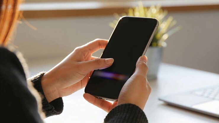 Mani di una donna con un maglione scuro che controllano il telefono alla scrivania vicino alla finestra, il pollice preme la parte inferiore dello schermo