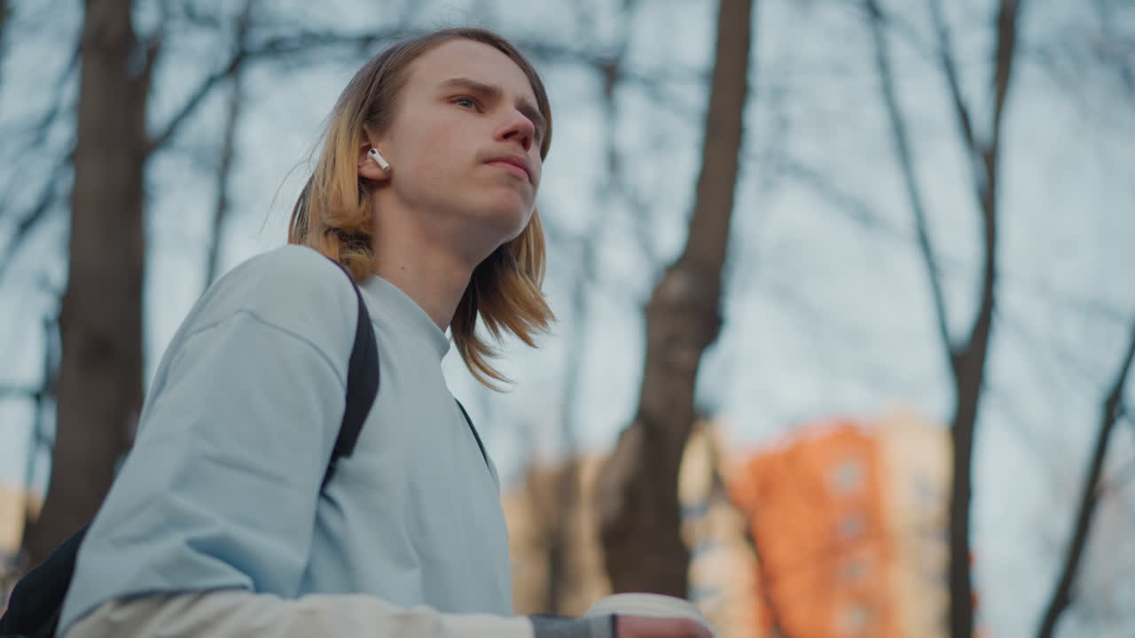 호기심 어린 표정으로 이른 봄 공원에서 커피를 마시며 주변을 둘러보는 흰색 옷을 입은 십 대 워커, 백팩과 이어폰이 보이고 도시 건물과 잎이 없는 나무가 장면을 둘러싸고 있어요.