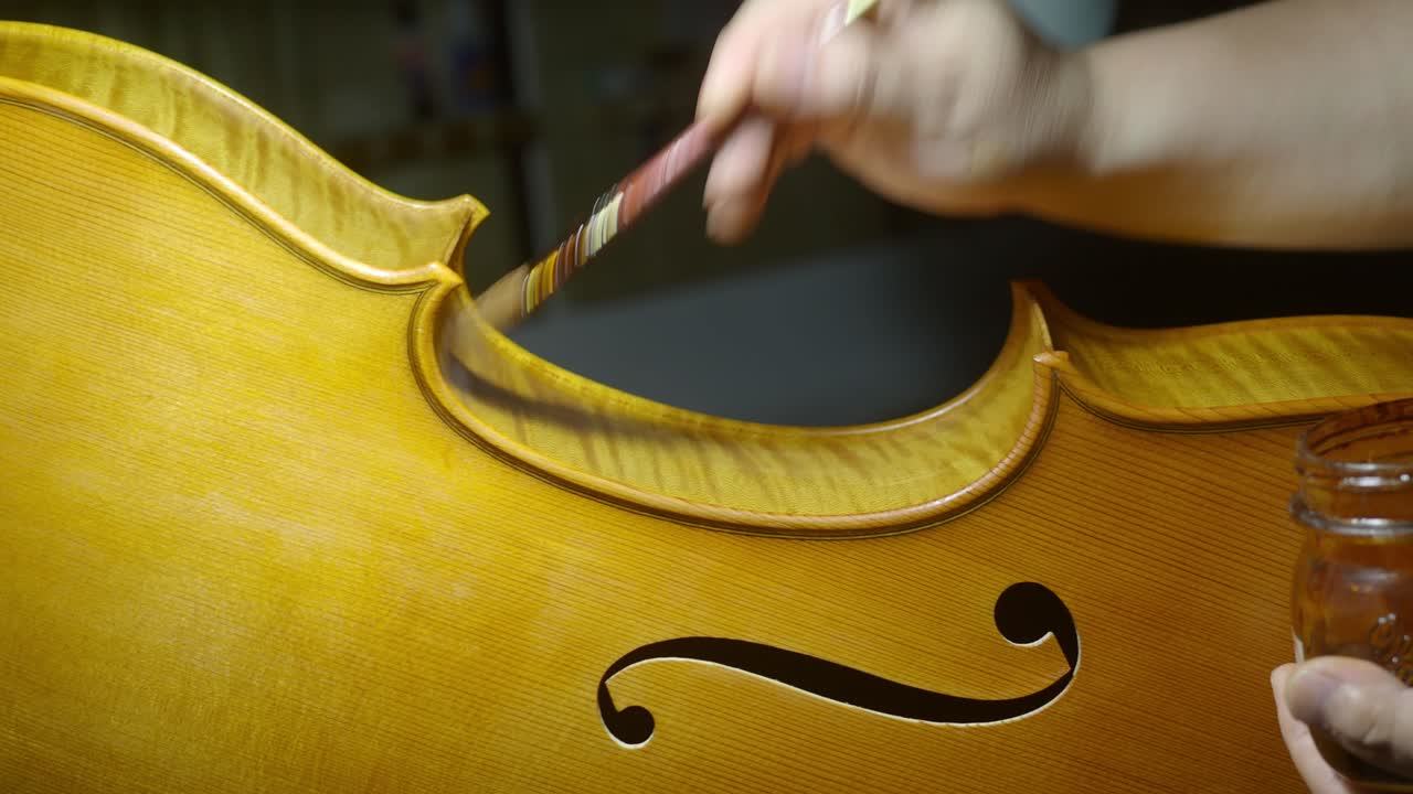 Luthier’s hand brushes natural blend of linseed oil, mastic, and lac along the ribs and curved edges beside the f-hole in the cello center bout, revealing detailed grain and craftsmanship, real time