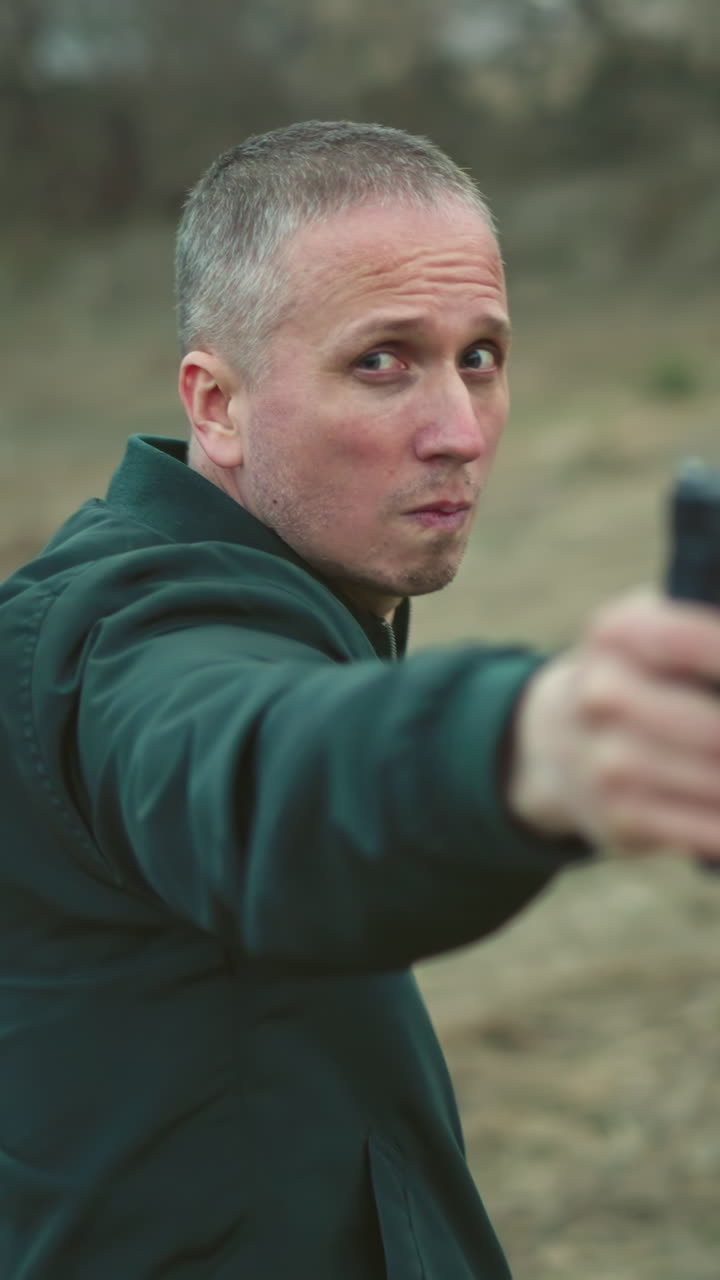 una vista cercana de un hombre con una chaqueta verde apuntando a una pistola mientras se da la vuelta en un entorno al aire libre cubierto de hierba
