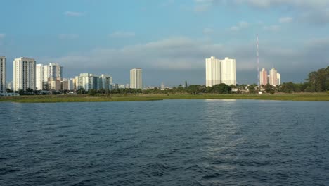 Aerial-drone-dolly-in-shot-of-the-Socorro-neighborhood-in-the-south-of-São-Paulo,-Brazil-from-the-man-made-Guarapiranga-Reservoir-with-calm-waters-and-skyscrapers-in-the-background-on-a-fall-evening