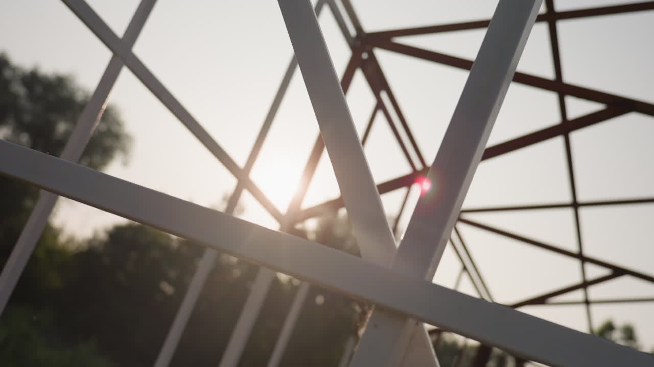 close up iron frame silhouetted against bright sky with sunlight flare and blur trees in background capturing geometric lattice pattern in soft backlight creating abstract architectural detail