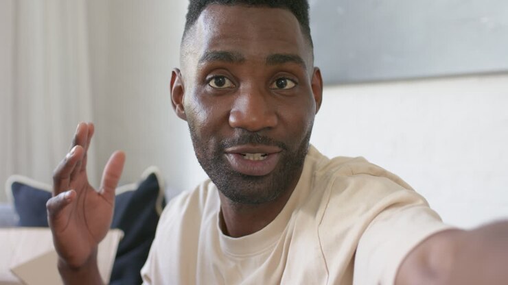 African American man placing hand on chest holding phone at arm's length on couch, sharing story