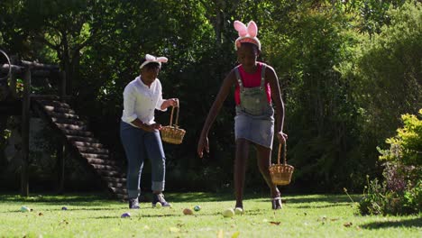 african american brother and sister wearing easter bunny ears doing easter egg hunt in garden