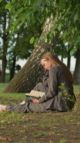 dama con ropa gris se sienta al aire libre bajo un árbol leyendo un libro con la mano en la página, la pierna estirada, la cabeza ligeramente inclinada mientras toca su cara el fondo presenta una vista borrosa de alguien caminando en la distancia
