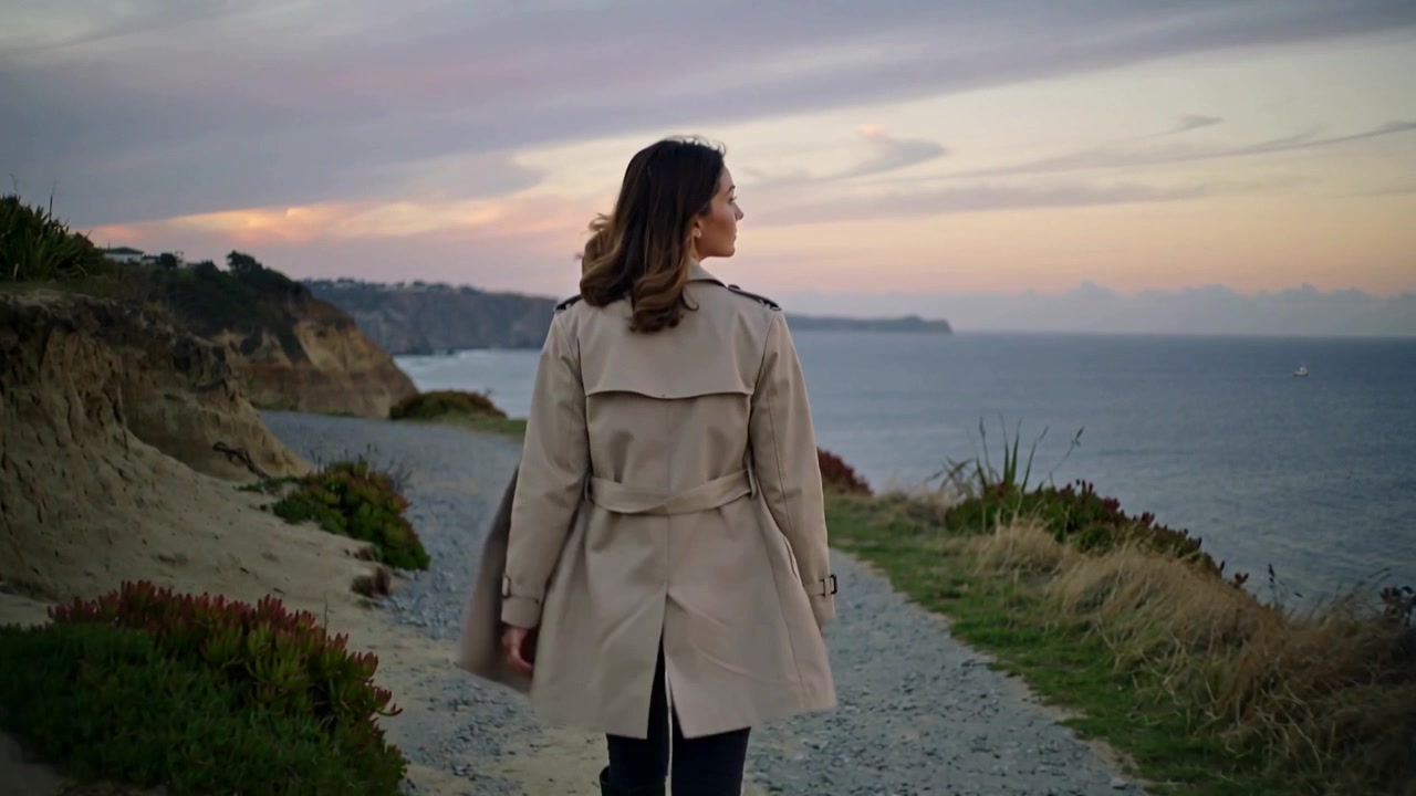 Walking woman in beige trench coat prompted by sunset, along gravel cliff path, glancing toward sea