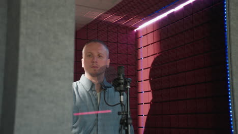 a man wearing a blue long sleeve shirt stands in front of a studio microphone in a red soundproofed room. the scene captures a moment in a professional recording environment