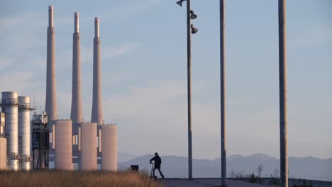 Ein-Mann,-Der-Eine-Industrielandschaft-Betrachtet,-Fährt-Bei-Sonnenuntergang-Mit-Einem-Elektroroller-Davon