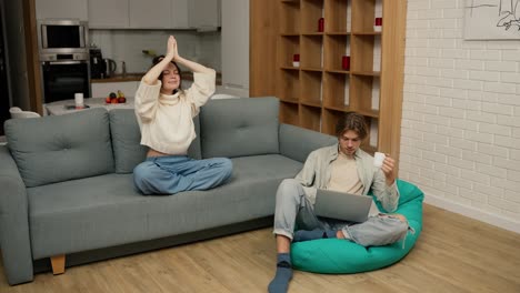 a woman doing yoga exercise or meditation while a man with notebook relaxing in soft chair with laptop