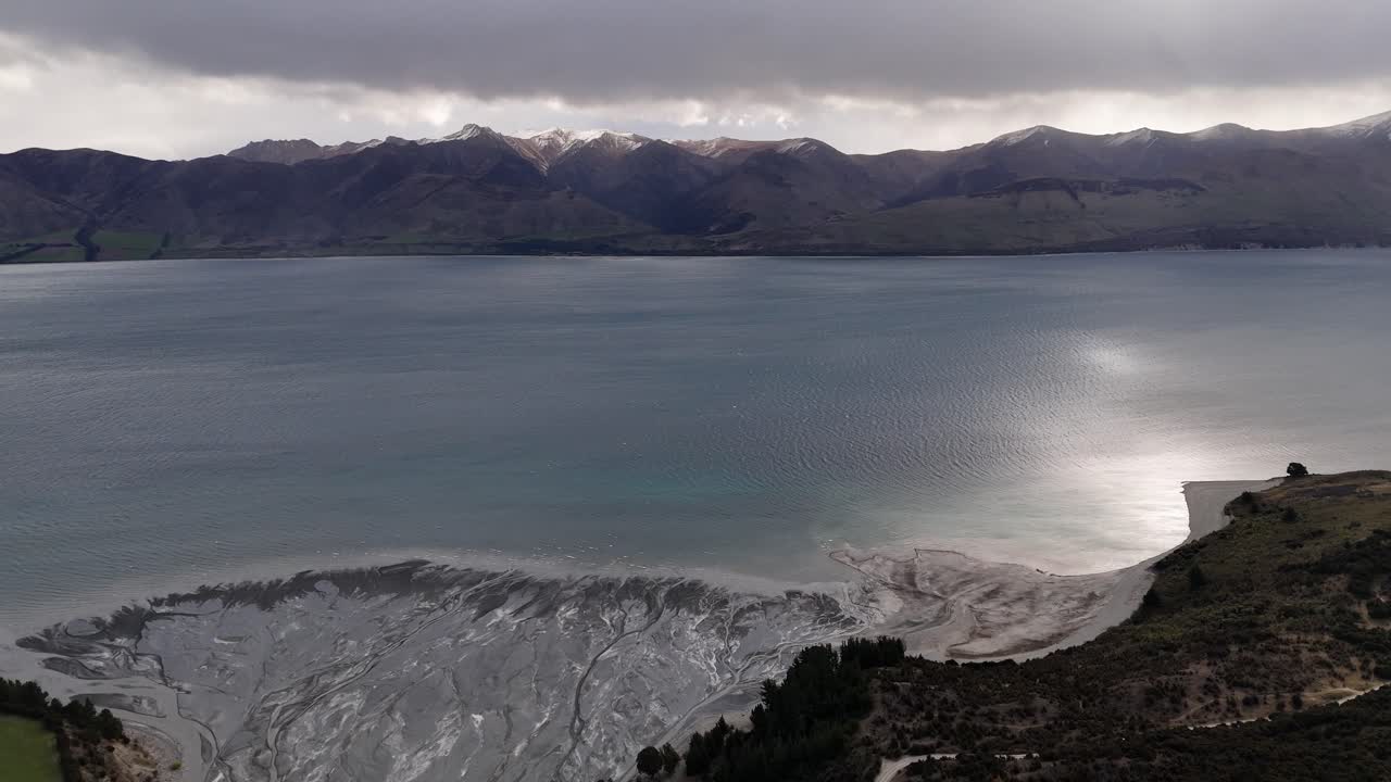 New Zealand Lake Hawea with dramatic mountains, reflective water, rugged shoreline, cloudy sky and textured river delta patterns. Calm natural landscape in NZ. aerial wide shot