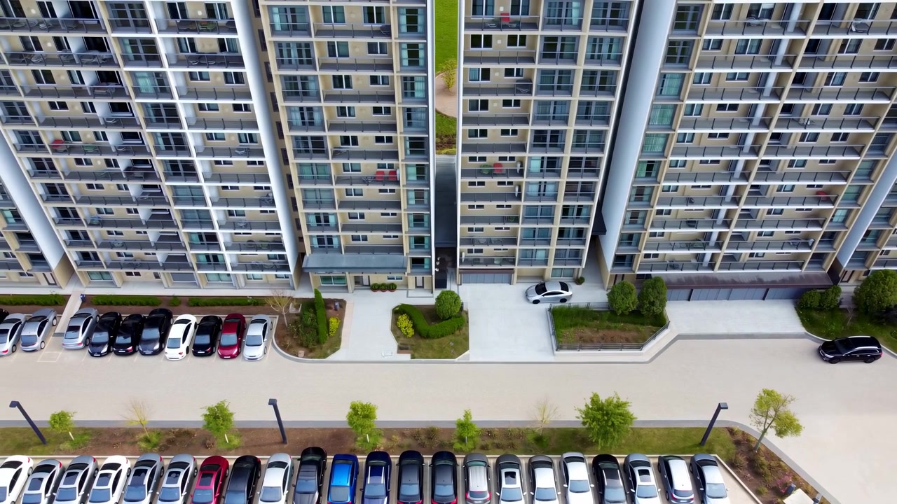 Pemandangan bergerak dari ketinggian di atas tiga menara apartemen yang berdekatan dengan mobil-mobil yang diparkir, kisi-kisi balkon, pintu kaca, pagar, kolom, pintu masuk beraspal, tepi lanskap, dan tata letak perumahan perkotaan yang tenang