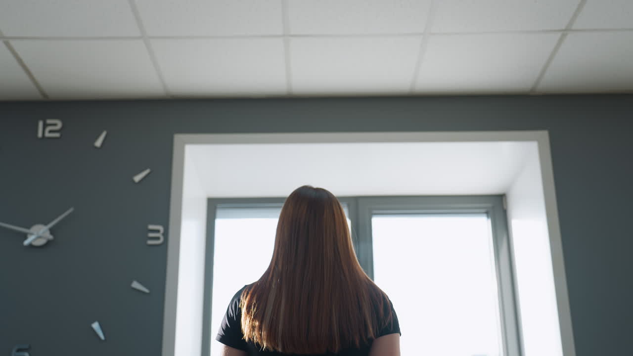 back view of woman with brown hair cascading around shoulders walking on treadmill facing wall clock and large window in modern gym studio under soft natural light and minimalist gray interior