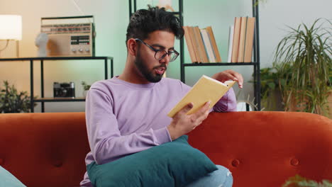 hombre indio leyendo un libro interesante volteando páginas sonriendo disfrutando de la literatura descansando en el sofá