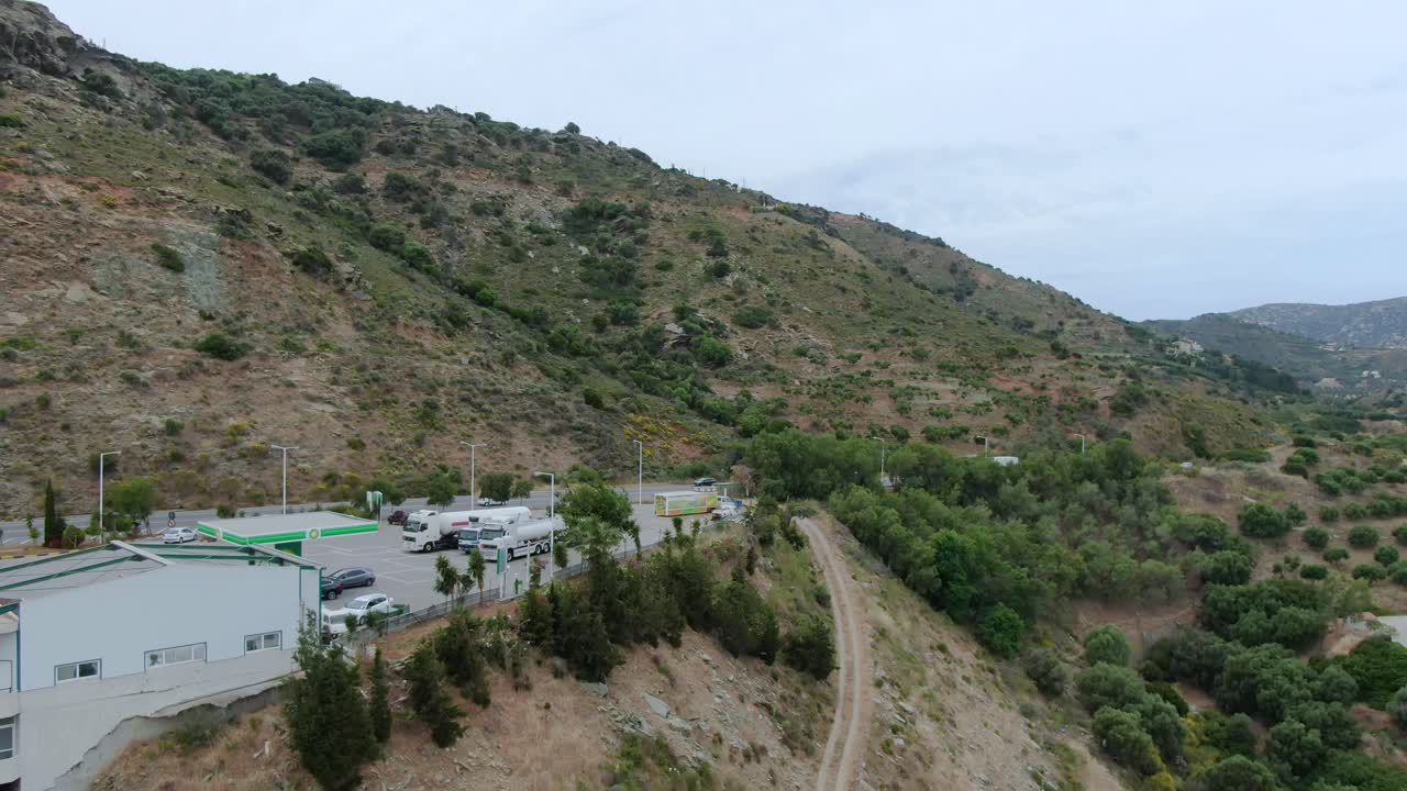 Aerial Over Petrol Station On Hillside In Crete With Cars Going Past. Follow Shot