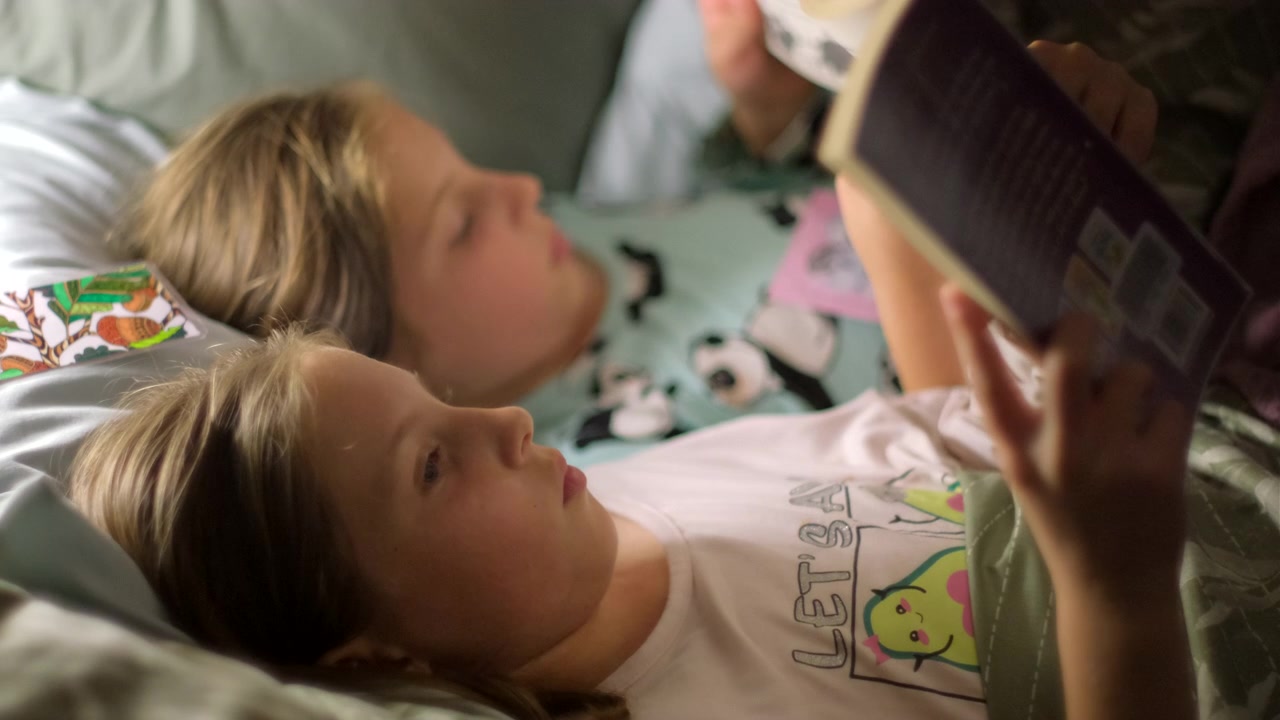 Twin sisters lying in pajamas together in bed reading bedtime stories at night