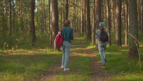 cousins walk along a scenic forest trail surrounded by lush greenery, one playfully hangs her black bag over her back while the other in a green shirt watch her