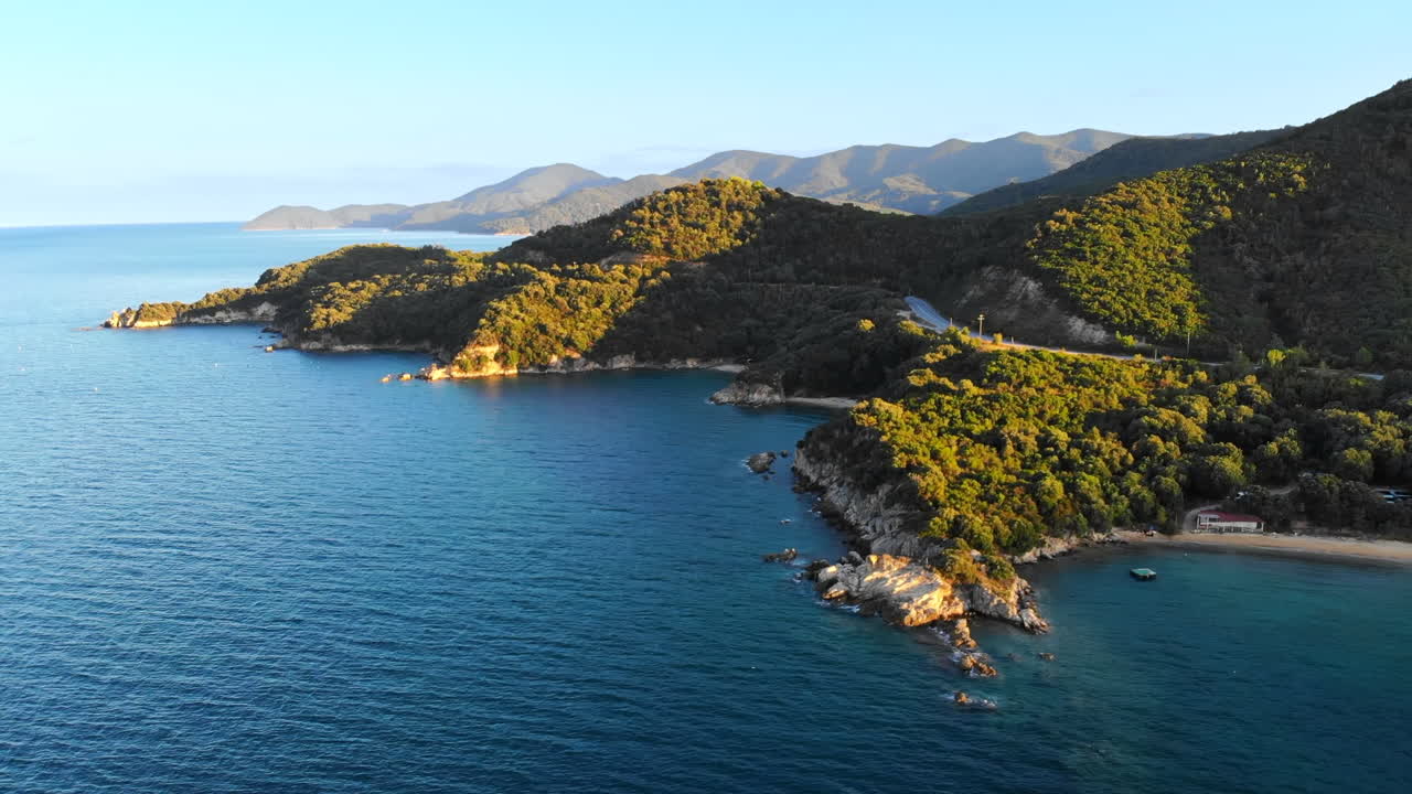 Aerial drone view of the Aegean sea rocky coast with greenery and road on the shore, hills in the distance in Greece