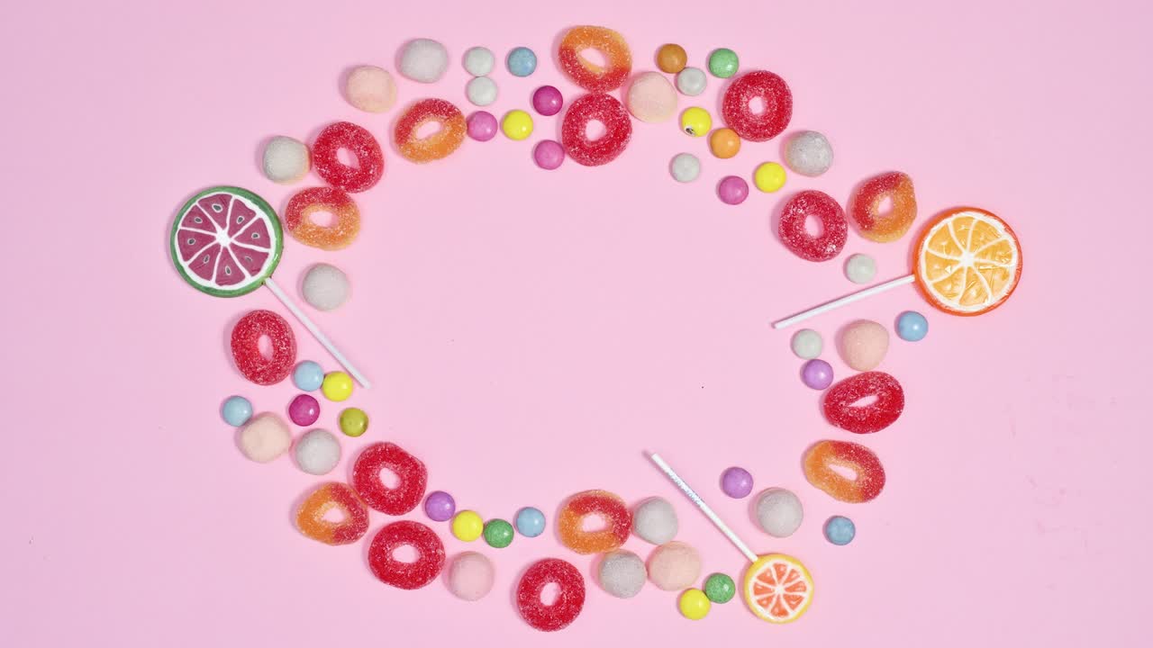 Sweet gummy candies, chocolate candies and lollypops appear in circle making frame on pastel pink background. Stop motion animation flat lay