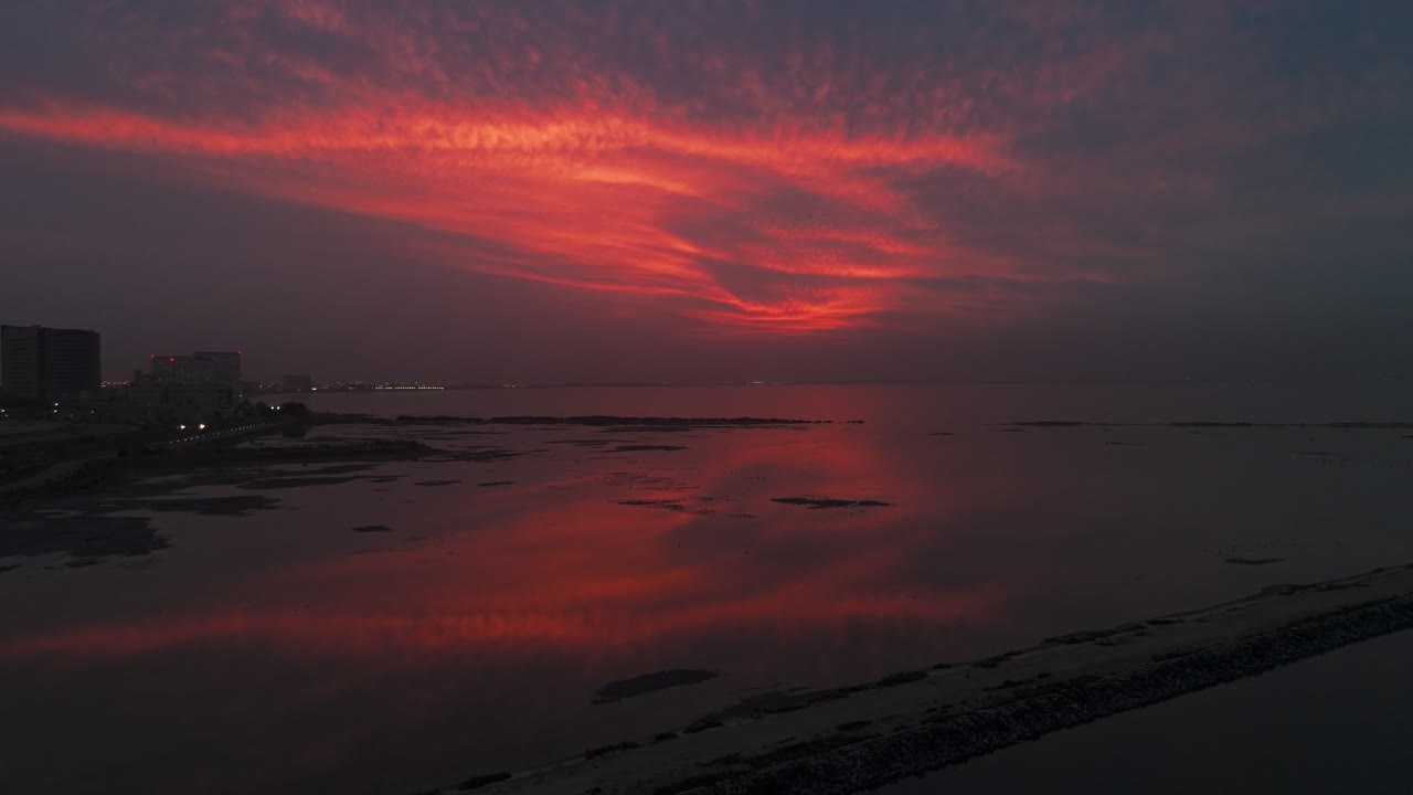Wide view of a dramatic red sunset sky reflecting over calm coastal waters, with distant city skyline silhouettes and shoreline visible under evening light