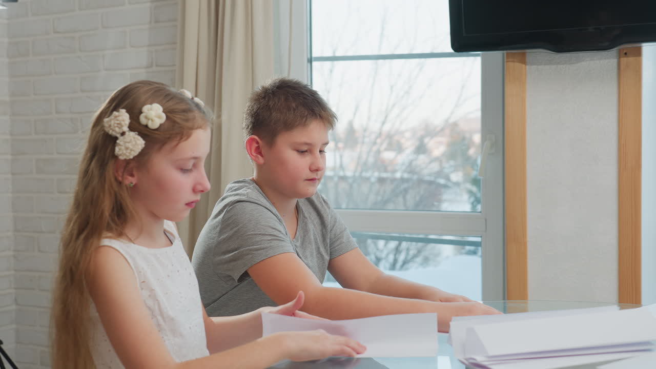 Kids in casual wear creating paper craft at table with winter weather background visible through window, snow outside with television in the background. Focus on paper crafting activity