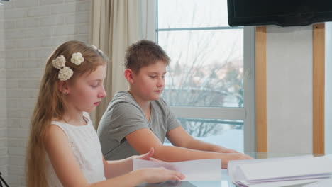 kids in casual wear creating paper craft at table with winter weather background visible through window, snow outside with television in the background. focus on paper crafting activity