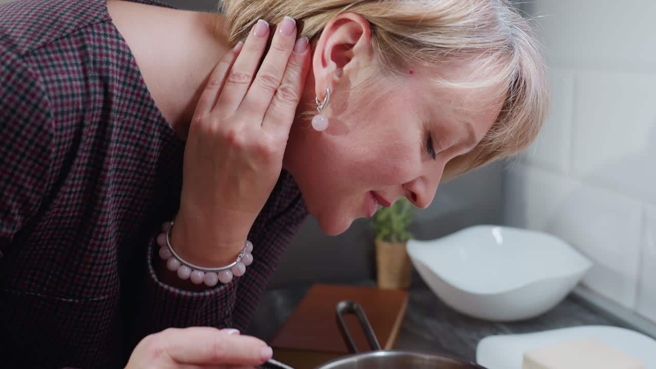 Mulher de cabelo loiro curto cozinhando em uma cozinha moderna, inclinando-se para sentir o aroma da comida com um sorriso satisfeito, mão no pescoço adornado com uma pulseira de contas rosa, segurando uma concha e tofu em um prato