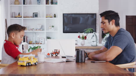 Hispanic-father-and-son-working-opposite-each-other-at-dining-table,-side-view,-waist-up,-lockdown