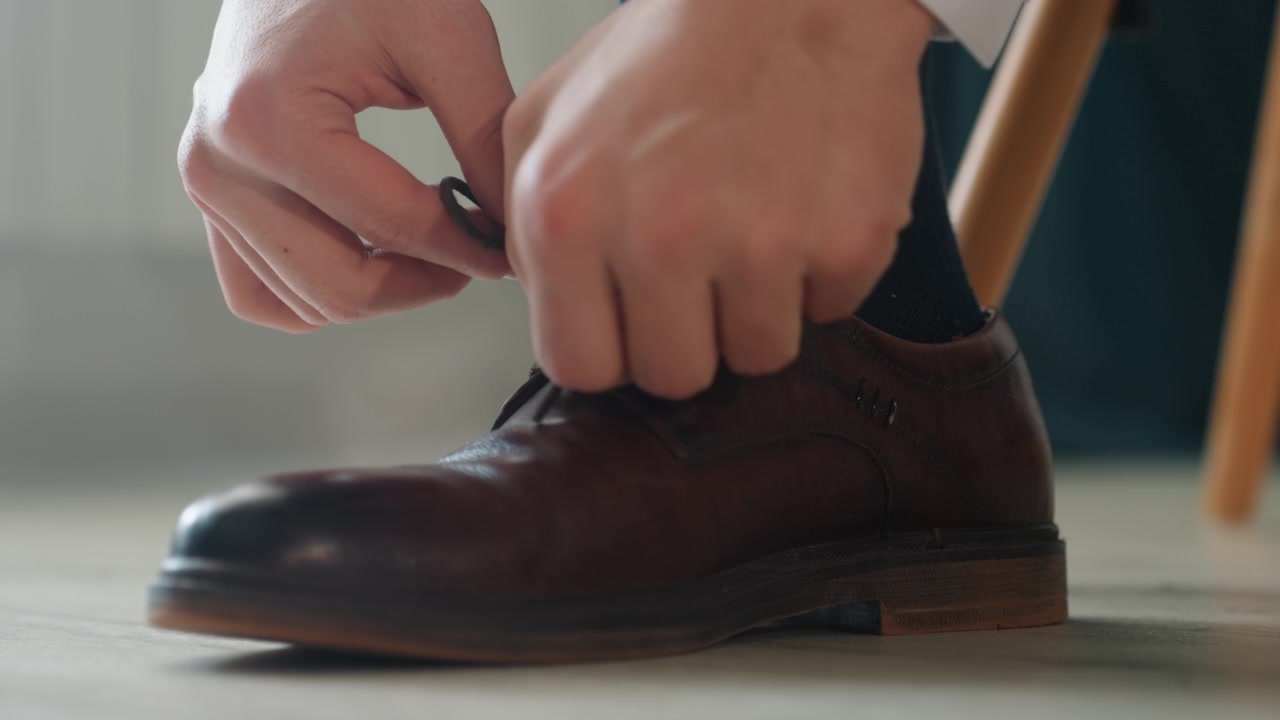 Intimate closeup brown shoe being tied, calm morning preparation with hands fastening leather oxford on wooden floor near chair polished detail, classic brogue texture, slow cinematic motion conveying
