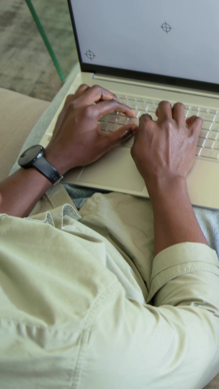 Vertical video: Typing after laptop showing crosshairs, man shifting hands on sofa in green shirt