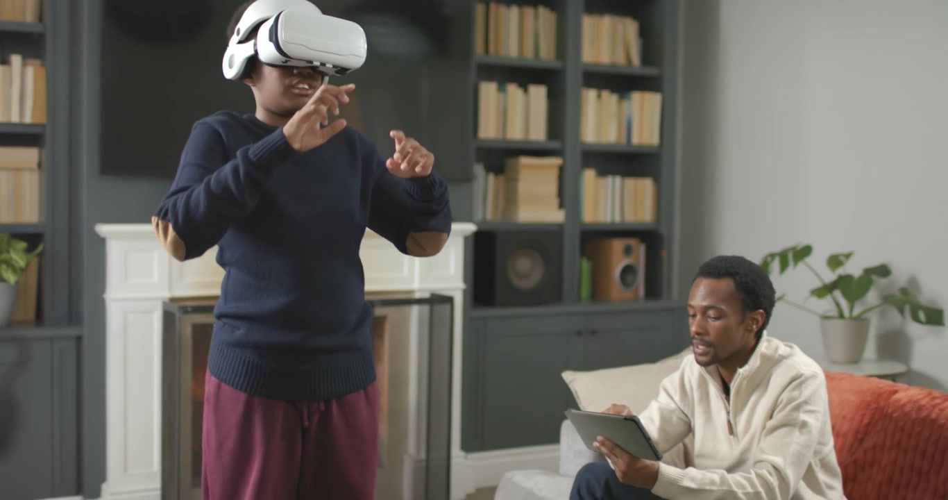 Male friends, one putting on white VR headset, interacting in VR at home, other tapping tablet