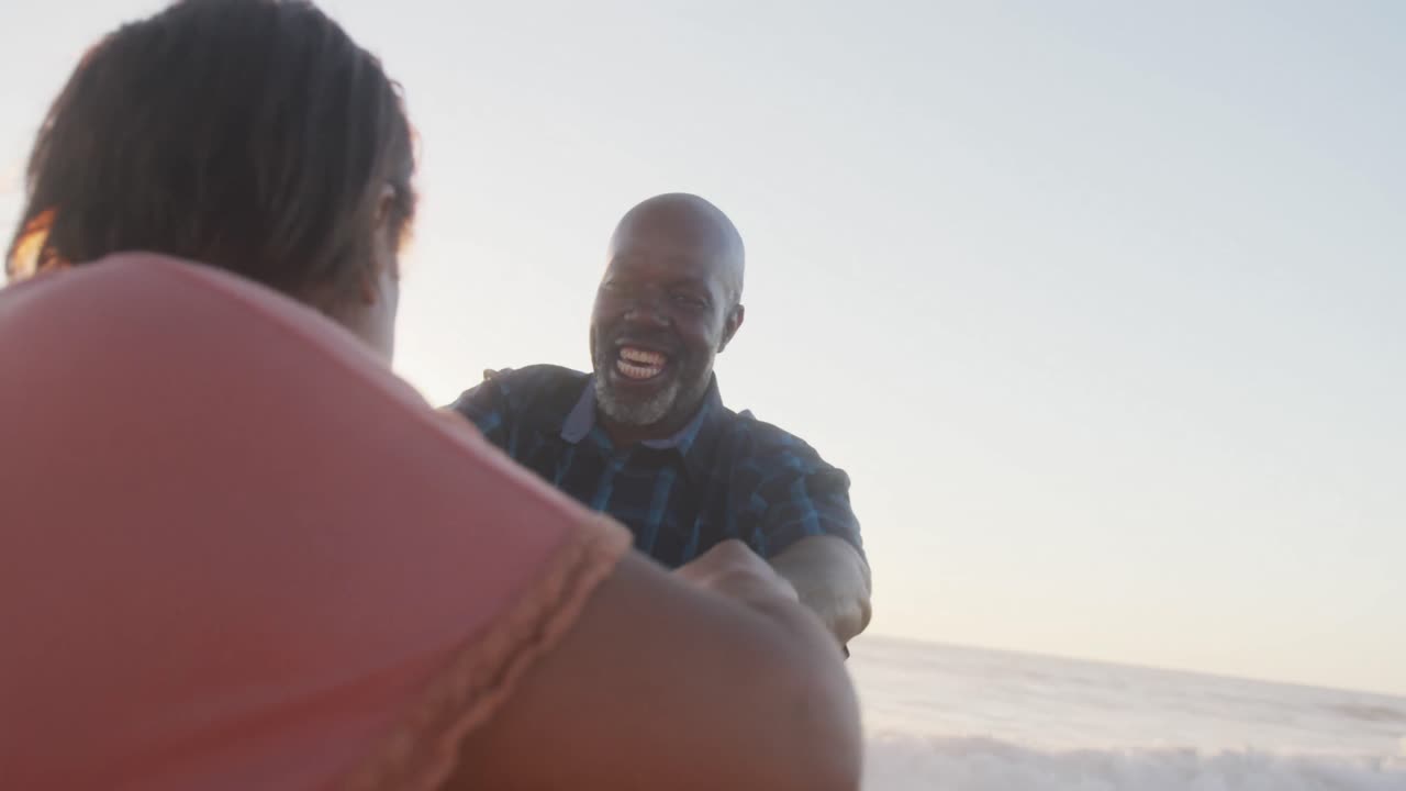 Smiling senior african american couple holding hands and dancing on sunny beach