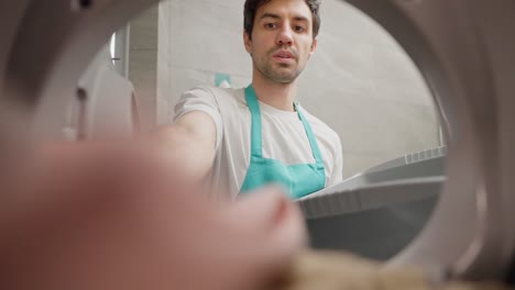 A-confident-brunette-janitor-guy-in-a-white-T-shirt-and-a-blue-apron-puts-dirty-clothes-from-a-gray-plastic-basin-into-the-washing-machine-and-starts-it