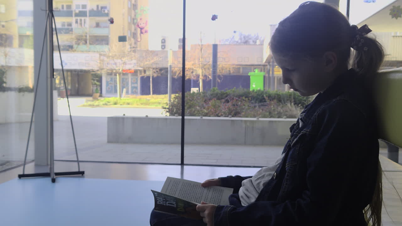 Profile view of a girl reading a book by a large glass window. Natural color grade and sharp lifestyle textures in a 2026 style