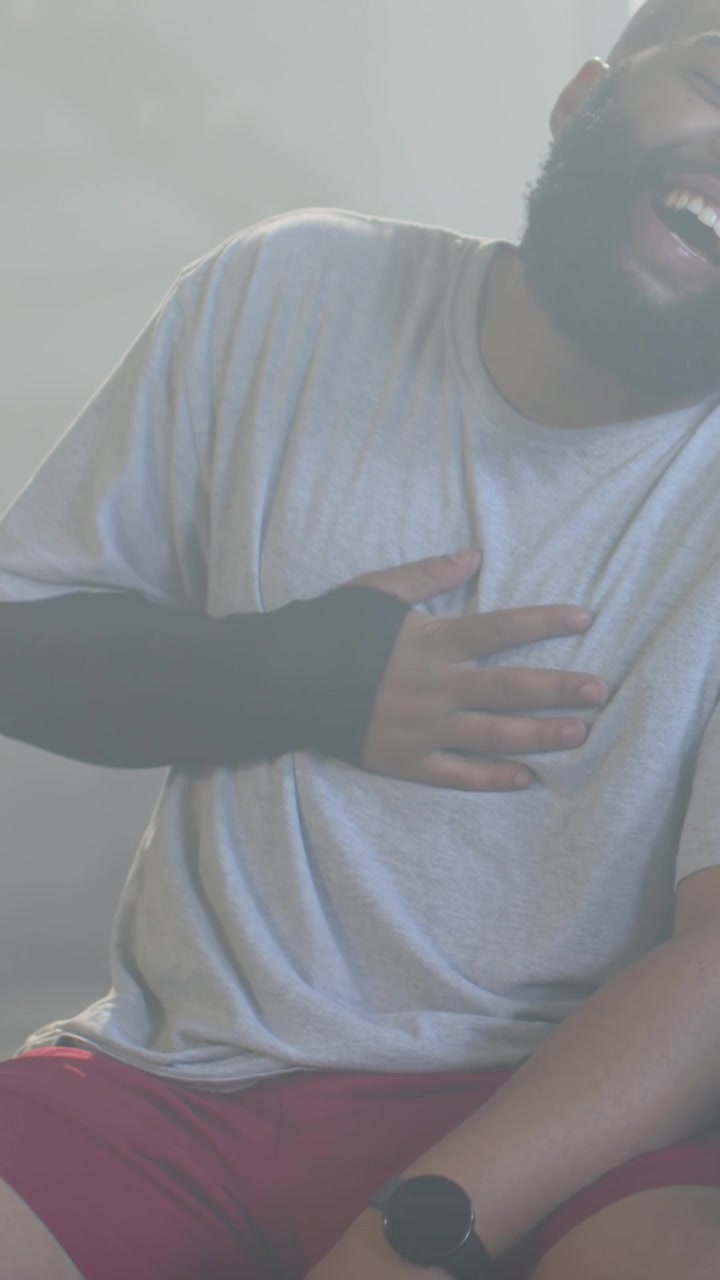 Vertical video: Laughing man reacting to gloved hand on chest in backlit room, red shorts and watch