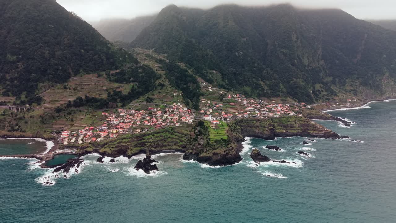 Vista aérea con dron de un dramático y oscuro pueblo costero de Madeira en un promontorio rocoso rodeado de olas oceánicas tormentosas.