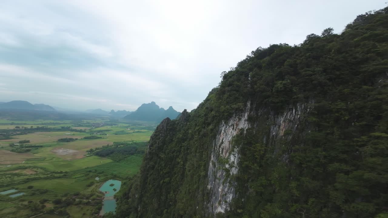 Aerial drone view of the Blue Lagoon, Laos, showcasing green fields and misty mountain peaks under cloudy skies with steep drop off cliffs