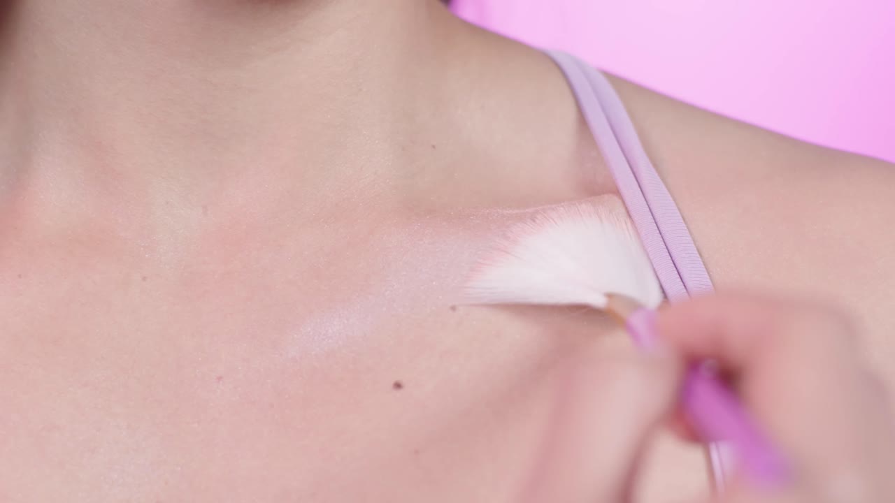 Close up of a young woman applying highlighter on her collarbone with a fan brush, make up, cosmetics, on a pink background