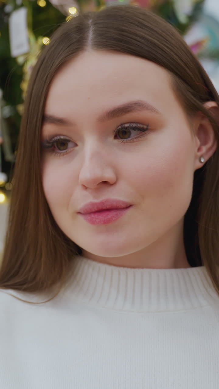 Happy lady in white sweater smiles while taking a picture in front of vibrant Christmas tree and colorful ornaments at a well-lit holiday decor store