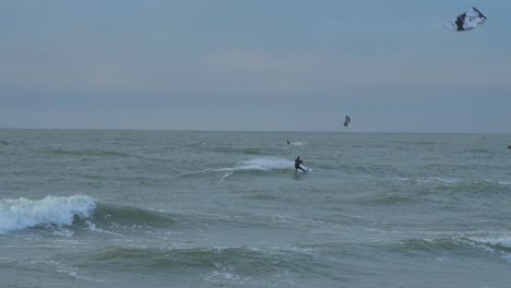 Eine-Gruppe-Von-Menschen,-Die-Kitesurfen,-Bewölkter-Wintertag,-Hohe-Wellen,-Ostsee-karosta-strand,-Zeitlupe,-Weitschuss