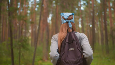 back view of young woman hiking through lush forest, wearing backpack and blue bandana, hands holding backpack straps, surrounded by tall trees and vibrant greenery