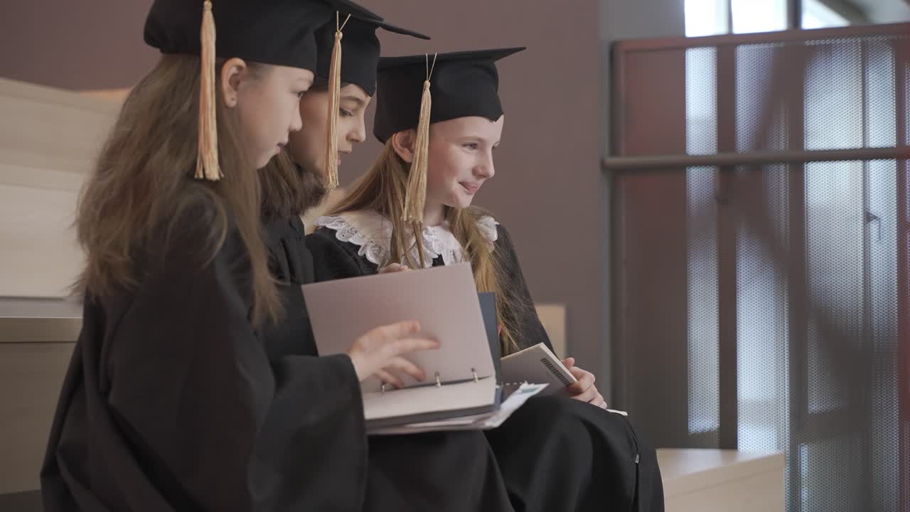 vista lateral de tres niñas felices con toga y birrete hablando y mirando sus cuadernos mientras se sientan en las escaleras en la ceremonia de graduación preescolar