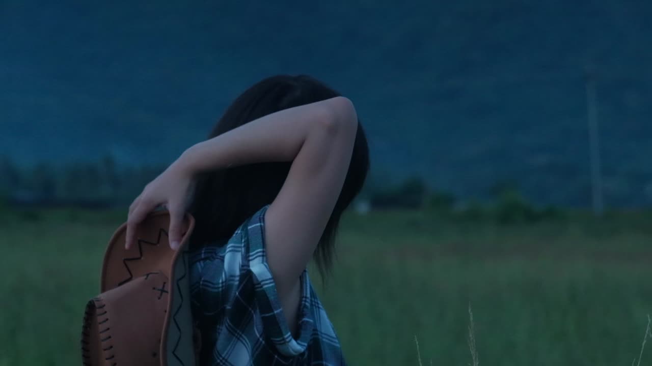 Rear static shot of black-haired cowgirl in flannel shirt reaching for cowboy hat during overcast day - slow-motion