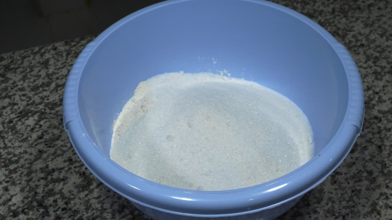 Mano di un fornaio che lascia cadere semi di anice in una ciotola blu contenente farina di grano e zucchero