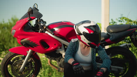 a female biker is seated on the roadside, legs crossed, touching her helmet in contemplation while sitting next to a red motorcycle