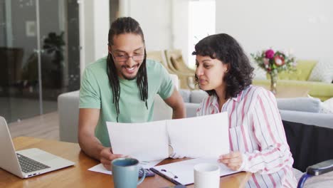 mujer biracial feliz en silla de ruedas y compañero masculino sonriente con papeleo y computadora portátil en la sala de estar