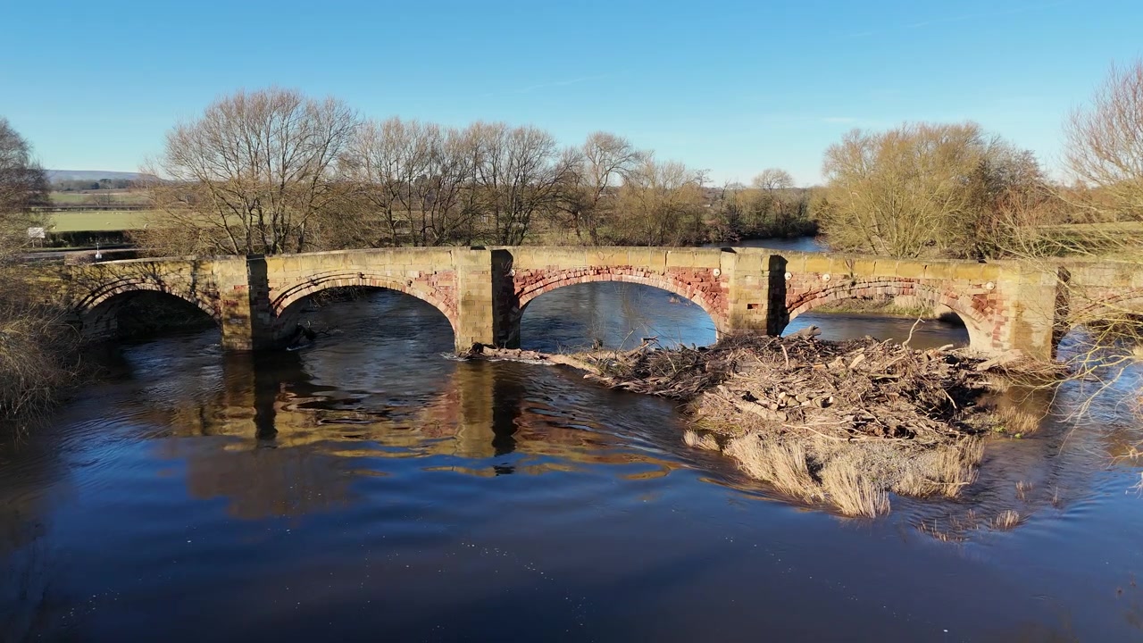 Ripresa aerea fluida con drone di un antico ponte ad arco in pietra e mattoni che attraversa un fiume in piena nella campagna inglese, con alberi invernali, paesaggio rurale e detriti naturali accumulati vicino al ponte