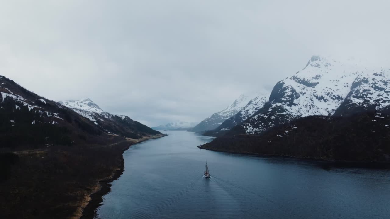 Vista aérea de veleiro entrando em fiorde norueguês, água azul calma entre montanhas cobertas de neve, paisagem nórdica dramática e conceito de aventura marítima ártica