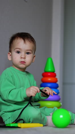 calm caucasian child sits on the floor playing with a black cord. balls, pyramid and big yellow toy box near the kid. vertical video